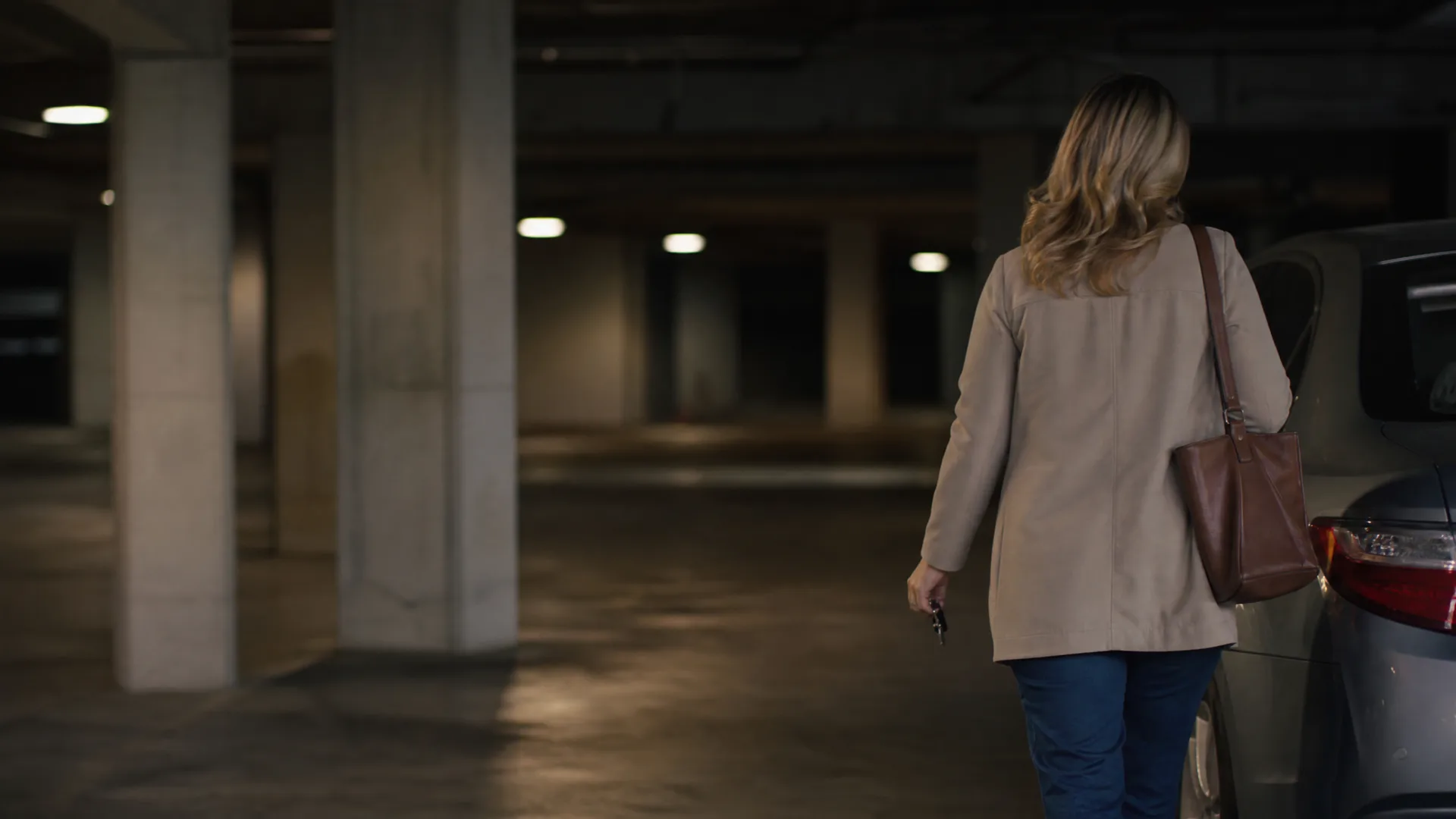 A blonde woman in a beige coat and jeans, carrying a brown shoulder bag and holding keys, walks away from the camera toward a parked car in a dimly lit concrete parking garage.