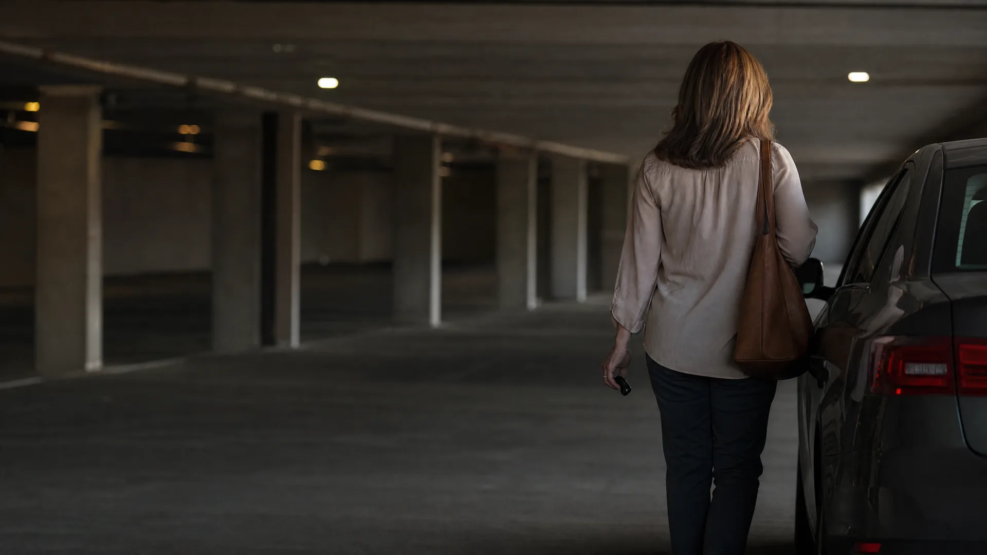 A woman seen from behind, carrying a brown shoulder bag and holding a key fob, walks toward a dark car in a dimly lit concrete parking garage.