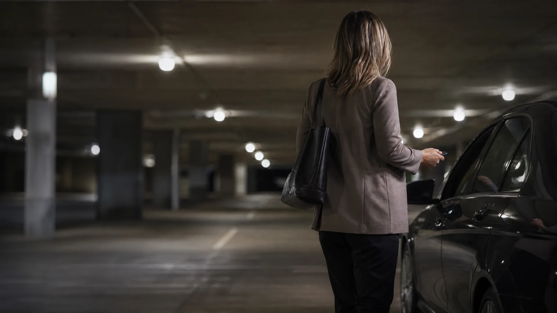 A woman seen from behind, wearing a light blazer and dark trousers with a black shoulder bag, stands beside a dark car in a dimly lit concrete parking garage.