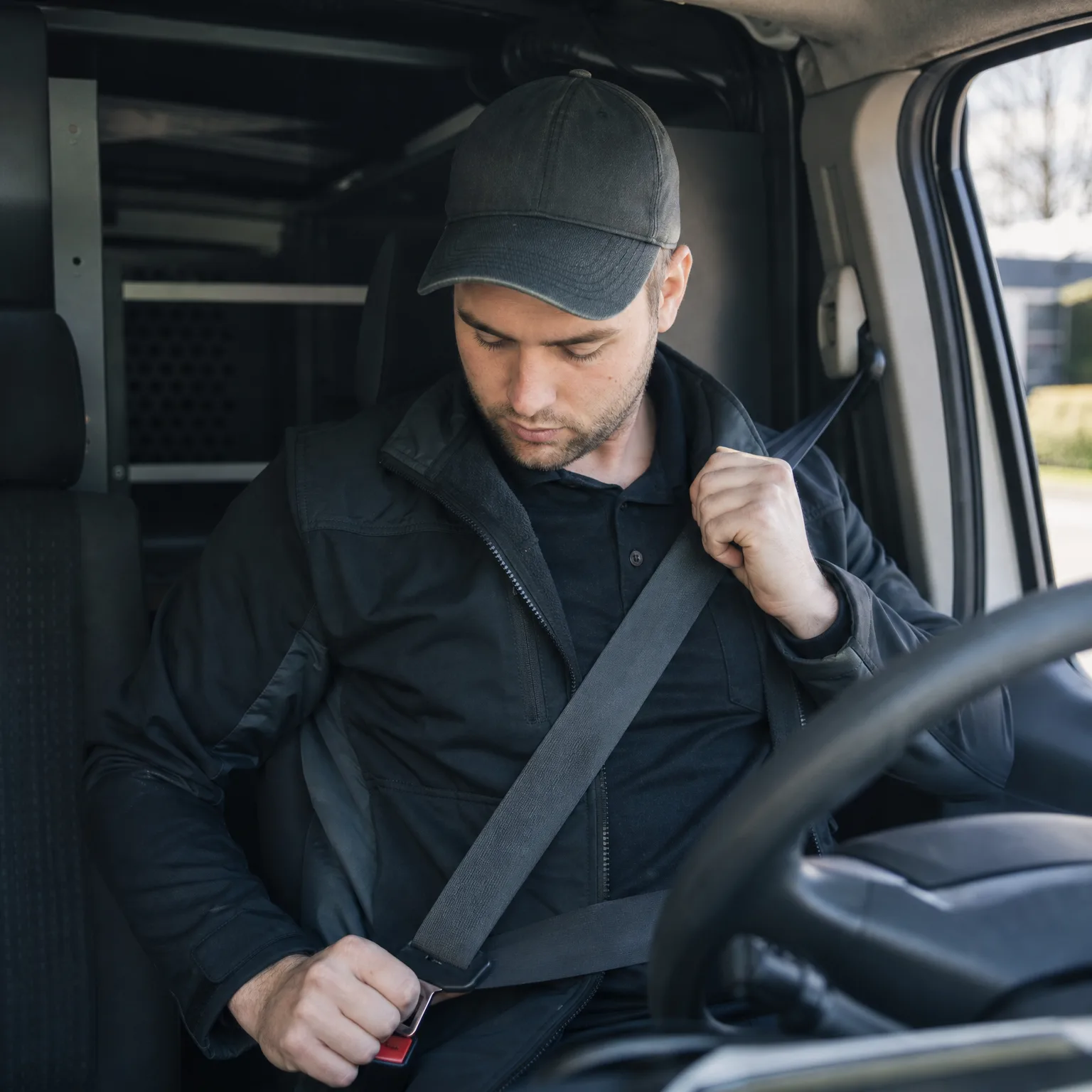 A man in a dark cap and dark jacket sits in a van's driver seat, fastening his seat belt with both hands, with the steering wheel at right and a window showing bare trees outside.
